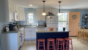 A kitchen with white cabinets, a central island with bar stools, a refrigerator, and a sink. - Salvo Sandbox: Ocean-Side, Dog-Friendly Cottage (Salvo)
