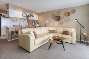 A modern living room with a beige sectional sofa, a small coffee table, and a kitchen area with white cabinets and a microwave.