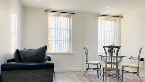 Een moderne woonkamer met een bank, een eettafel met stoelen en grote ramen met jaloezieën.