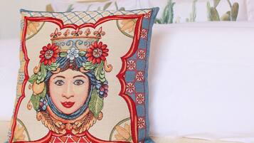 Un almohadón con un diseño bordado detallado, sobre una cama con ropa blanca y una planta de cactus al fondo.