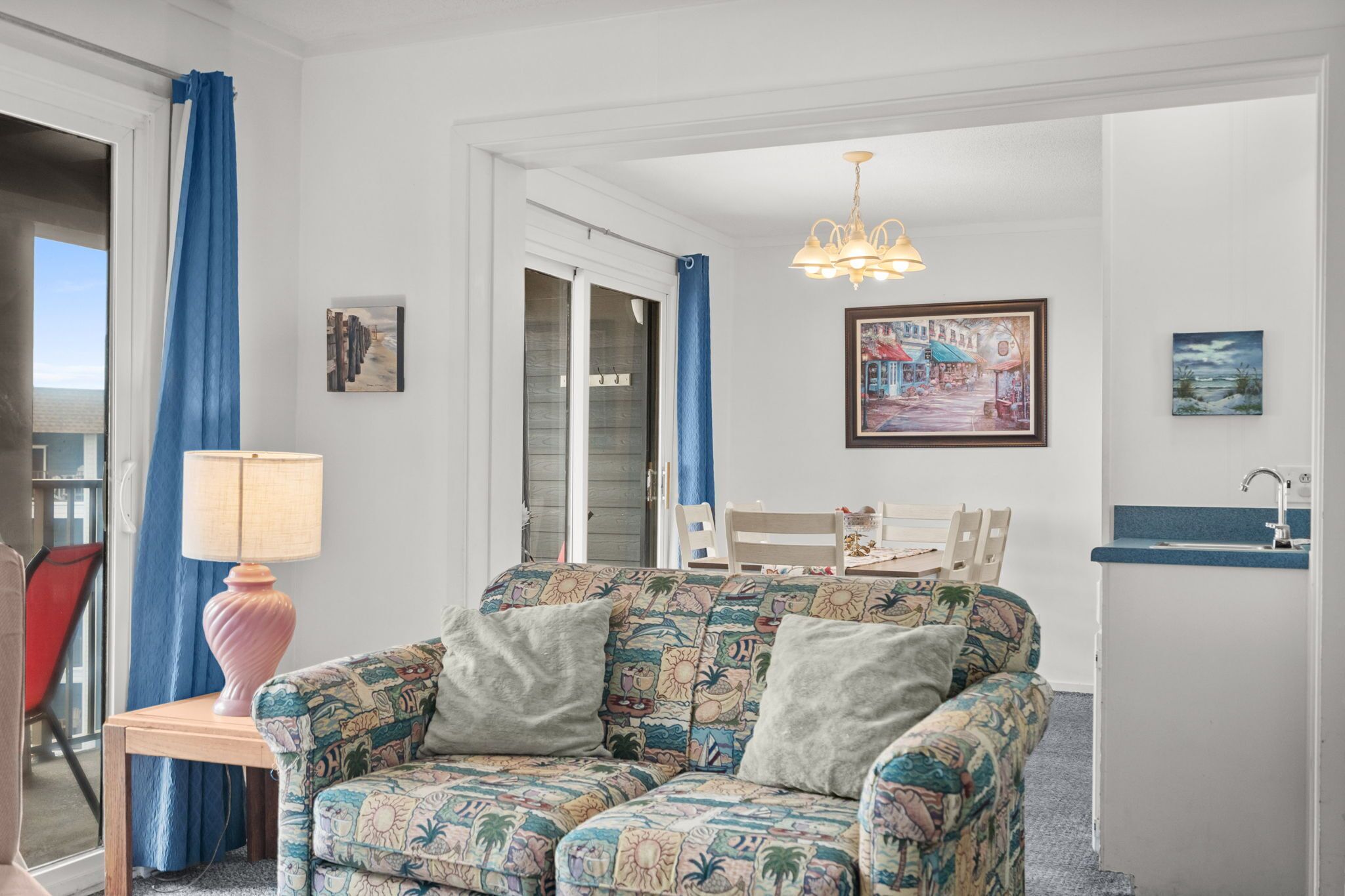 A living room with a patterned sofa, a table with a lamp, a chair, and artwork on the walls.