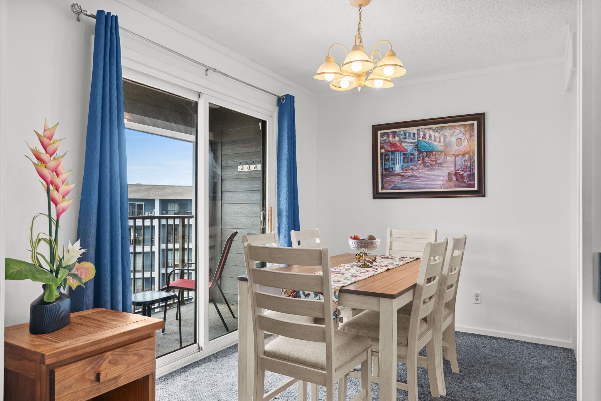 Uma sala de jantar com mesa, cadeiras, um candelabro, um quadro e uma varanda com cortinas azuis.