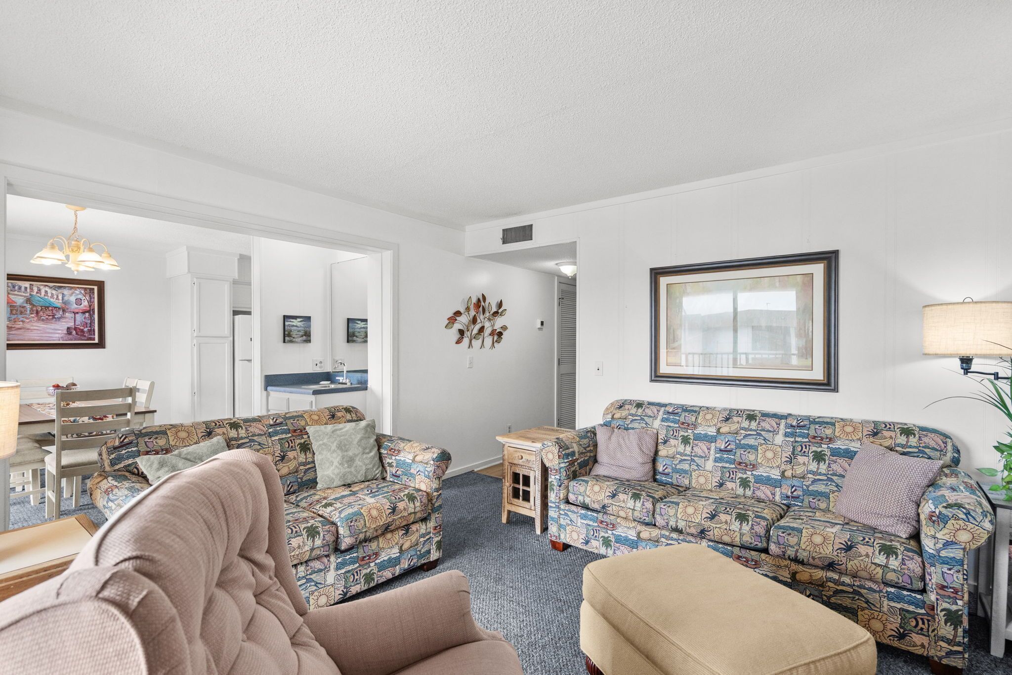 A living room with patterned sofas, a dining area, and a kitchen visible through an open doorway.