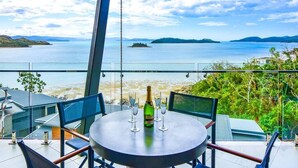 A round table with four chairs, a bottle of champagne, and two glasses on a balcony overlooking a body of water and distant islands.