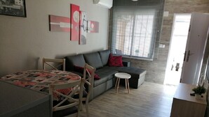Un espace de vie compact comprenant une table à manger, un canapé et un petit tabouret. On y trouve une fenêtre avec des stores, une porte et un réfrigérateur.