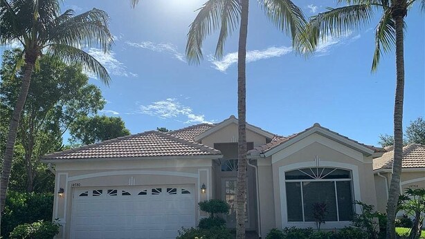 A single-story house with a white garage door, a covered patio, and a small front yard with palm trees and shrubs.