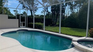 A swimming pool with a white tiled deck, surrounded by a covered area with a view of trees and a grassy lawn.