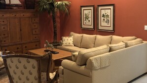 A living room with a beige sofa, a wooden coffee table, a patterned armchair, a dining table with chairs, a ceiling fan, and two framed botanical prints on the wall.