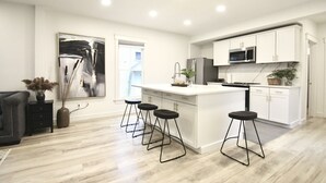 Une cuisine moderne avec des meubles blancs, un îlot central doté de tabourets, un four à micro-ondes et une grande fenêtre.