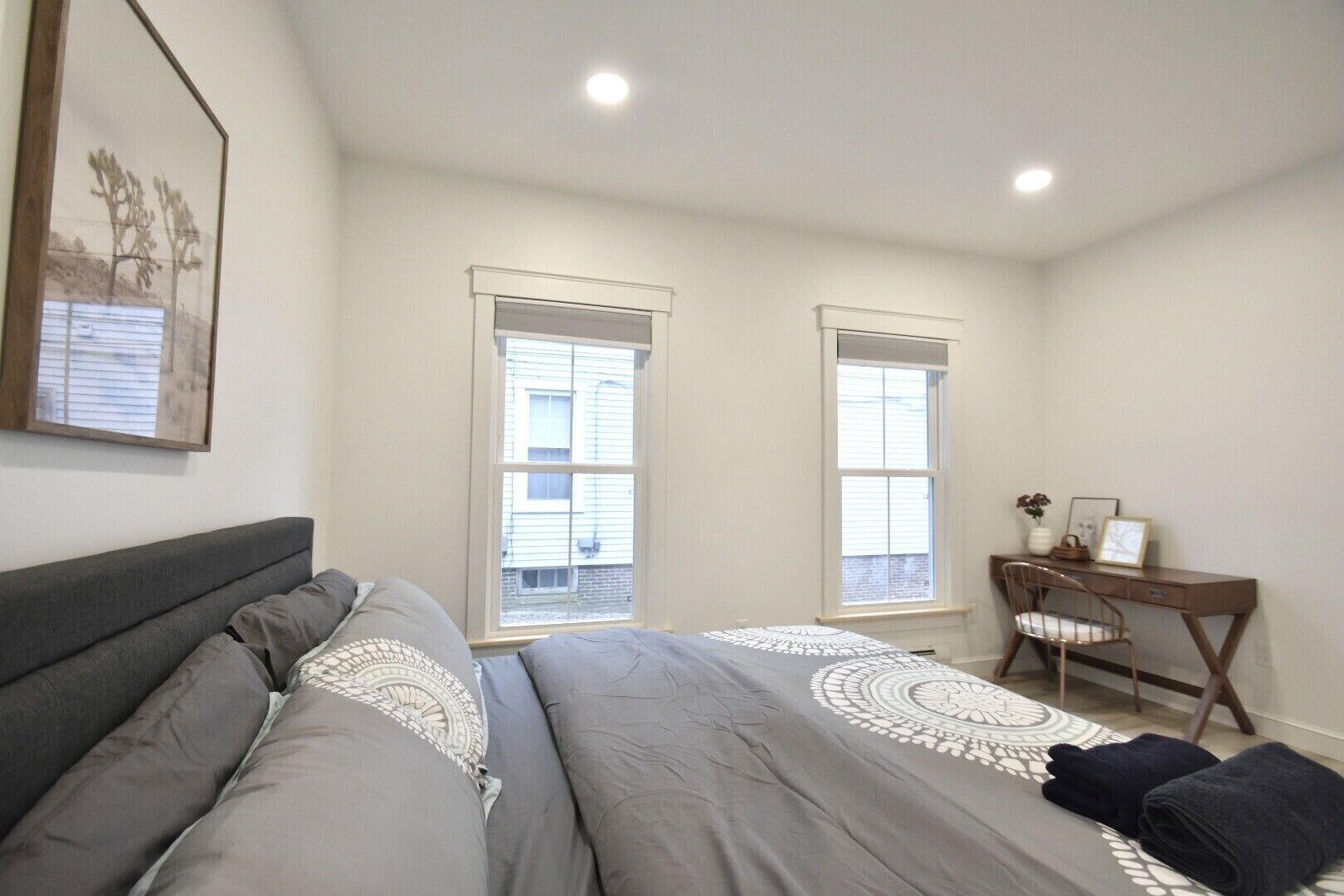 Ein Schlafzimmer mit zwei Fenstern, einem Bett mit grauer Bettwäsche, einem Schreibtisch mit Stuhl und einem gerahmten Bild an der Wand.