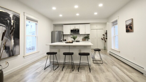 Une cuisine moderne avec des meubles blancs, un îlot central doté de tabourets, un réfrigérateur et un four à micro-ondes.