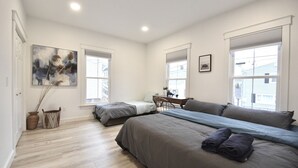 Une chambre avec deux lits, un bureau et une chaise. Il y a des fenêtres avec des stores, un tableau au mur et un vase contenant des plantes séchées.