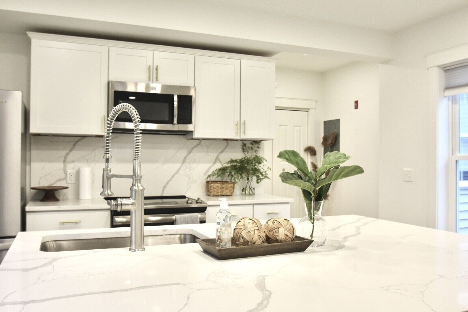 Una cucina moderna con pensili bianchi, lavello in acciaio inox, forno a microonde e una grande isola con piano di lavoro in marmo.