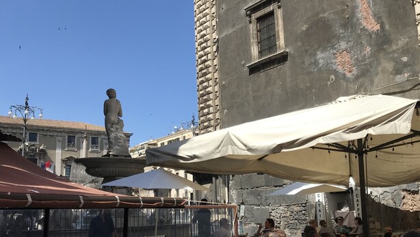 An outdoor seating area with tables and umbrellas, a statue, and a building with a stone facade. - Ade Home (Catania)
