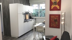 A compact kitchen with white cabinets, a gas stove, and a window overlooking greenery. A dining area with a white table and chairs is adjacent. - Ade Home (Catania)