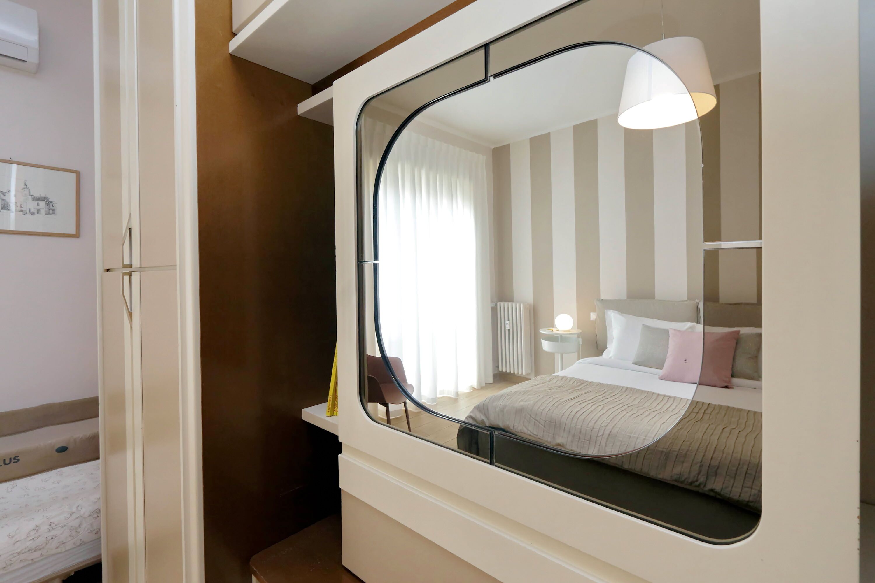 A bedroom with a large mirror, a bed with a striped bedspread, a bedside table with a lamp, and a window with curtains.