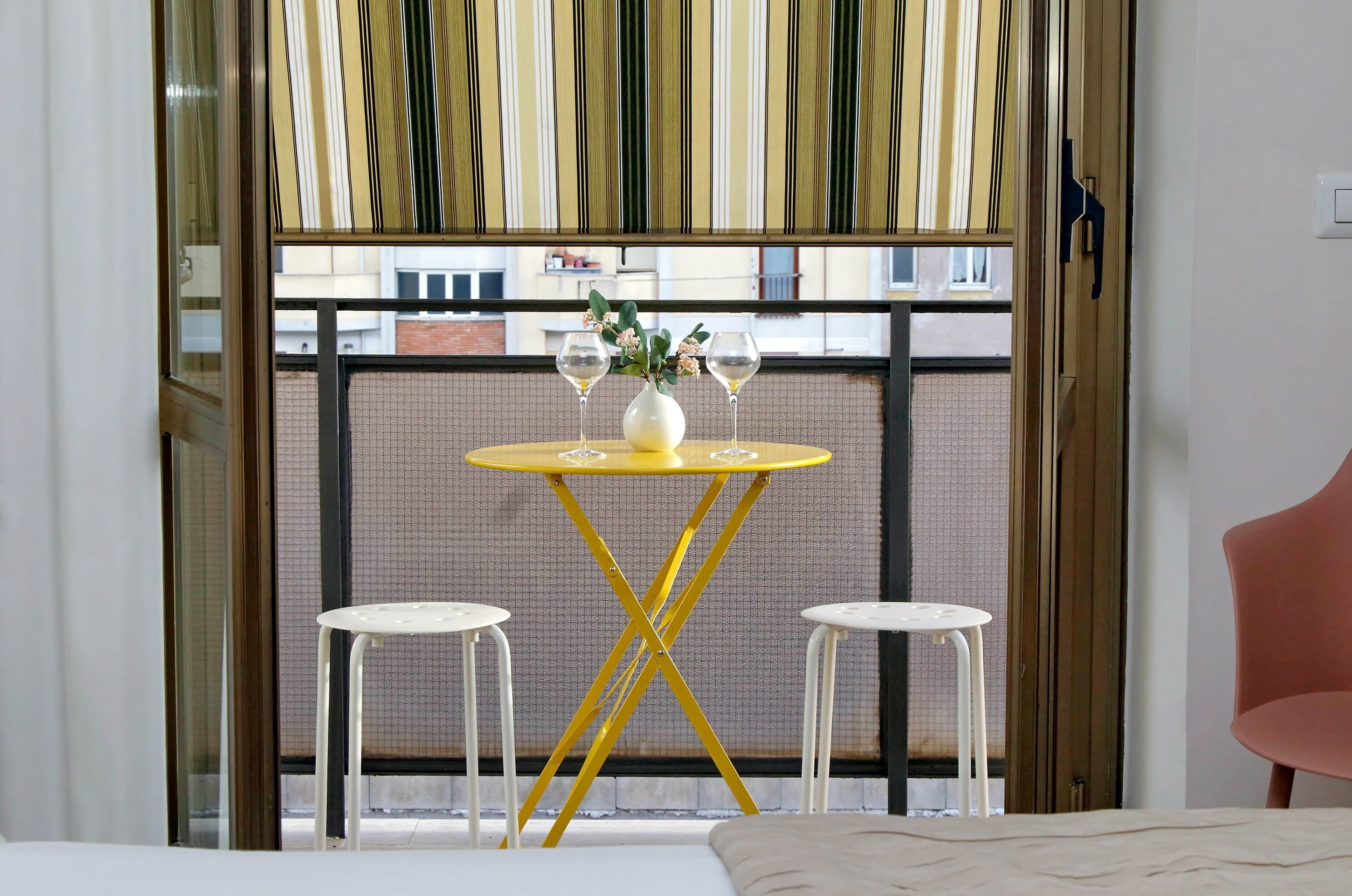 Een kamer met een gele tafel, twee witte stoelen, een bed en een raam met uitzicht op gebouwen.