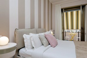 Una habitación de hotel con cama, almohadas, una mesita de noche con lámpara, una pequeña mesa con un florero y vistas a un balcón con cortinas.