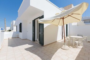 Ein weißes Gebäude mit einer gefliesten Terrasse, einem weißen Tisch mit Stühlen und einem beige gefärbten Sonnenschirm.