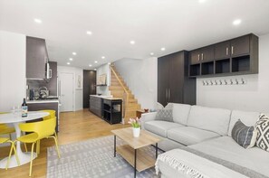 A modern living room with a sofa, coffee table, and yellow chairs. There is a staircase leading to a kitchen area with dark cabinets and a wooden floor. - Downtown Aspen Townhome—Minutes to Gondola/Art Museum (Aspen)
