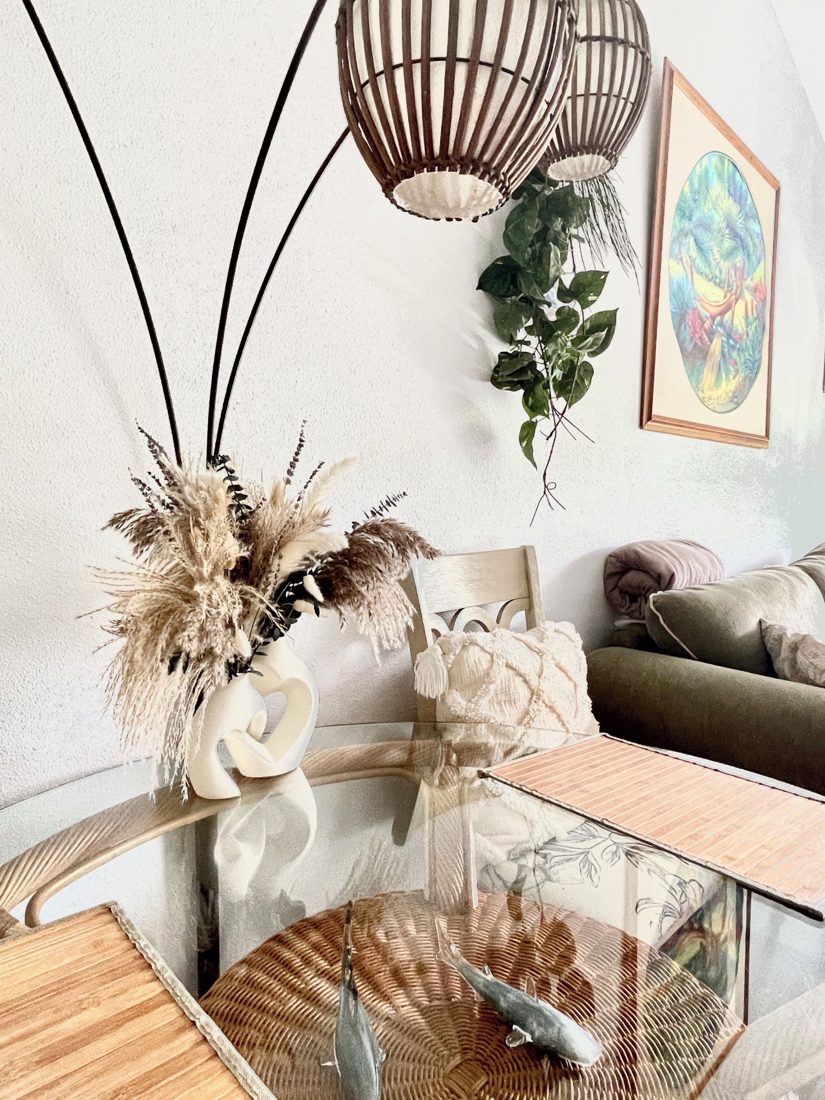 Ein modernes Wohnzimmer mit einem Glastisch, einer weißen Vase mit Trockenpflanzen, einem Korbstuhl, einem grünen Sofa, einem Holznachttisch und einer hängenden Leuchte.