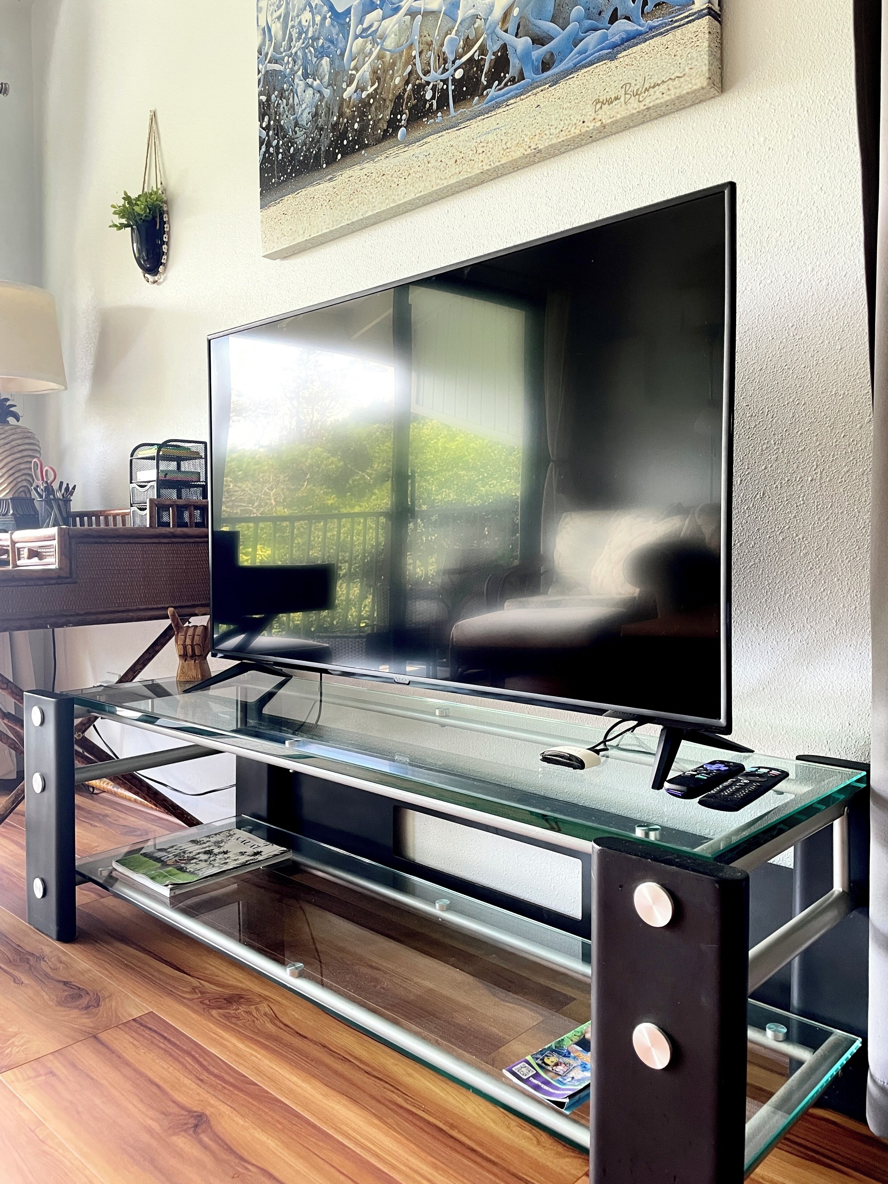 A modern living room with a flat-screen TV on a glass table, a bookshelf, and a painting on the wall.