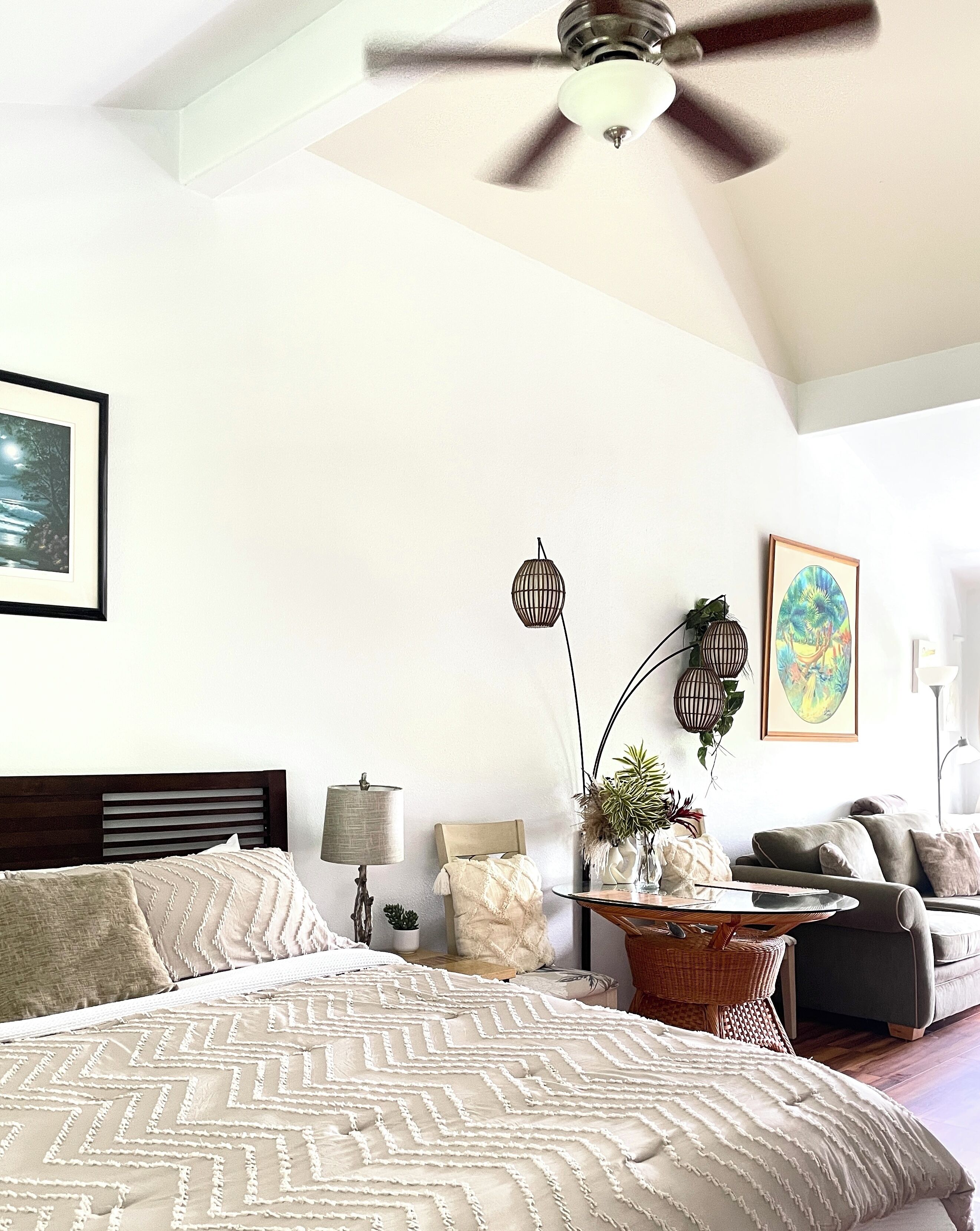 A bedroom with a bed, a ceiling fan, a nightstand, a lamp, a vase with plants, a painting, and a sofa.