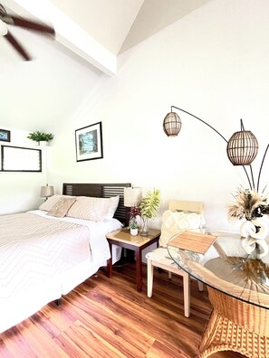 A well-lit bedroom with a large bed, bedside tables, a small table, and a chair. There are wall-mounted lamps and a ceiling fan.