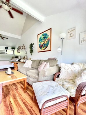 A living room with a wooden floor, a grey sofa, a wooden coffee table, a wicker chair, and a painting on the wall. - Tropical, Bright Princeville Studio with Mountain Views (Princeville)