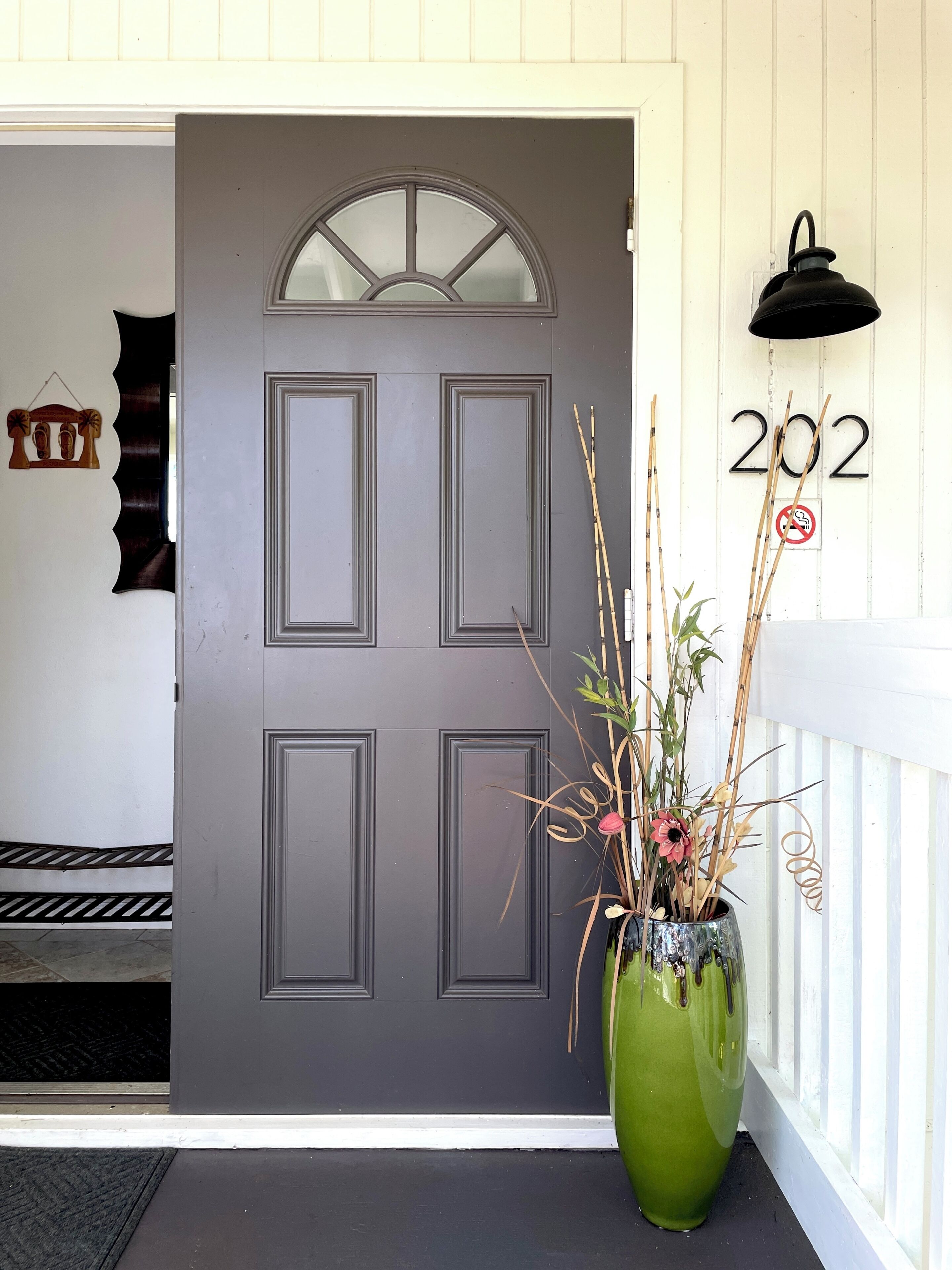 A grey door with a window above it, a green vase with plants on the porch, and a '202' sign with a no-touch symbol on the wall.