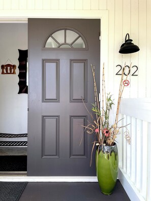 A grey door with a window above it, a green vase with plants on the porch, and a '202' sign with a no-touch symbol on the wall.