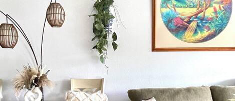 Un salon avec un plancher en bois, une table basse en verre, une table d’appoint en osier, un fauteuil rembourré, un canapé et une plante suspendue.