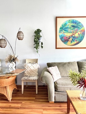 A living room with a wooden floor, a glass coffee table, a woven side table, a cushioned armchair, a sofa, and a hanging plant. - Tropical, Bright Princeville Studio with Mountain Views (Princeville)