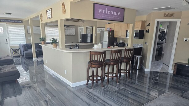 A modern living space with a kitchen area, a bar counter with stools, and a 'Welcome' sign. - BRAND NEW 16x14 Heated Swim Jacuzzi Sleeps 8 Beach Front! No Condo Park 4 Cars! (Fort Myers Beach)