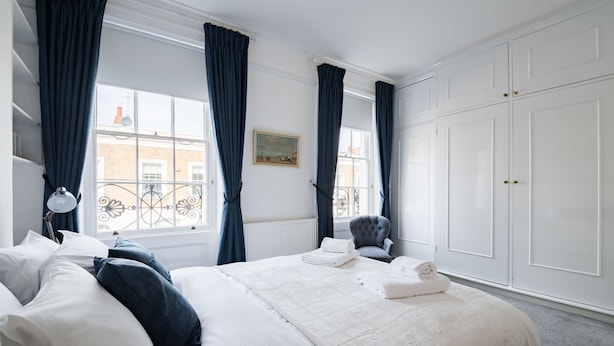 Una cama bien hecha con sábanas blancas y una silla gris, un armario blanco y una ventana con cortinas azules.