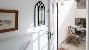 Un interior blanco con una puerta que da a un espacio exterior. Hay un cuadro en la pared, una ventana con marco negro y una mesa con sillas afuera.