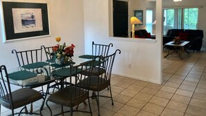 A dining area with a glass table, metal chairs, and a chandelier. There is a framed picture on the wall and a window with curtains. - Three Bedroom Condo - 8841 GP - Vusa (Kissimmee)