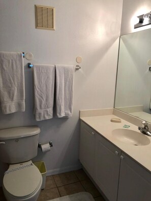 A bathroom with a toilet, two towels hanging on a towel rack, a sink with a mirror, and a window with blinds.