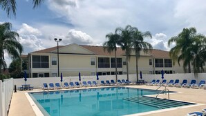 A pool area with blue lounge chairs and umbrellas, a clear pool, and a building with balconies in the background. - Three Bedroom Condo - 8841 GP - Vusa (Kissimmee)
