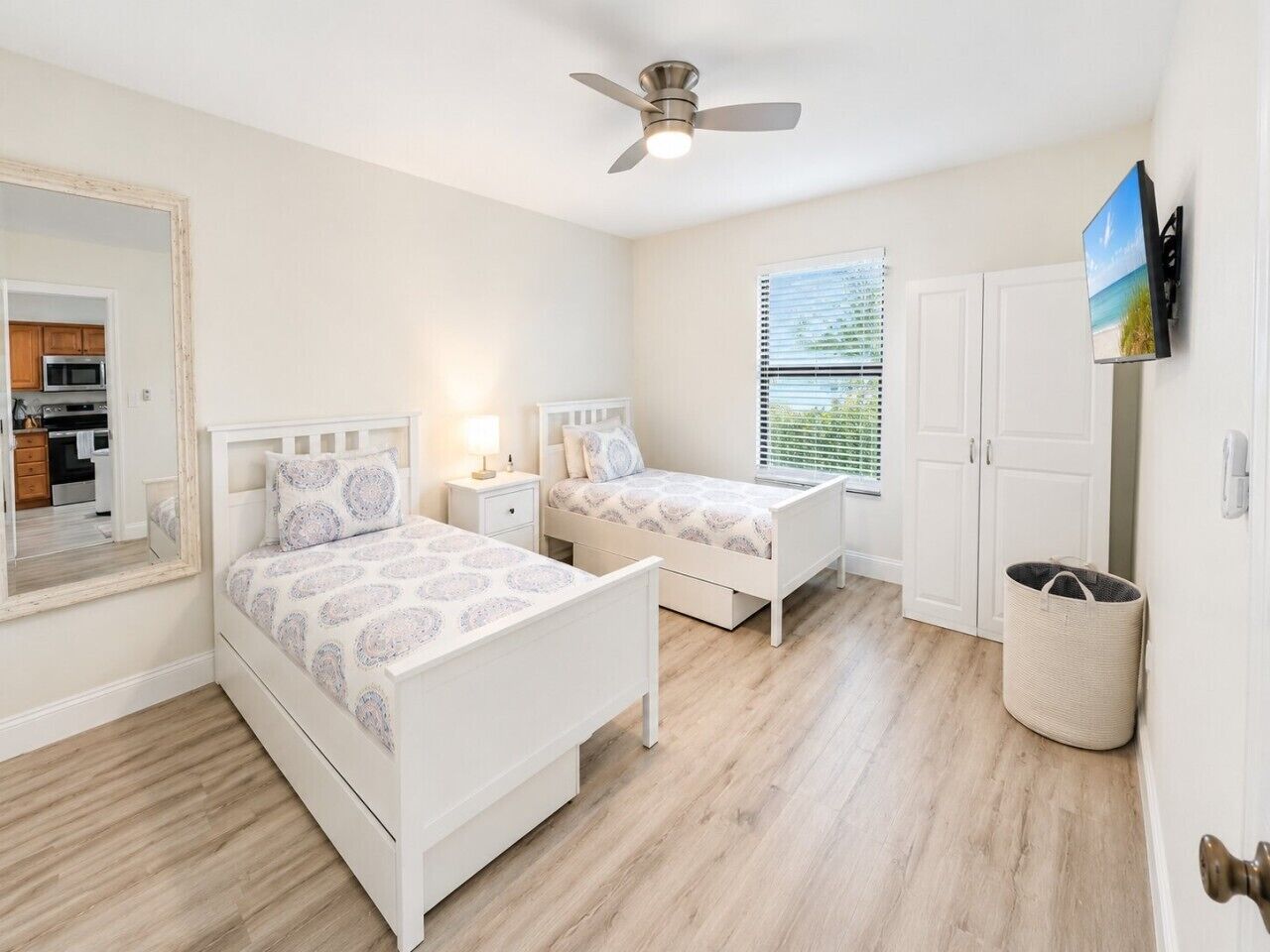A bedroom with two beds, a ceiling fan, a window with blinds, a TV mounted on the wall, and a laundry basket.
