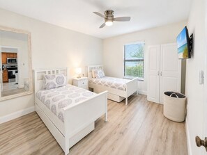 A bedroom with two beds, a ceiling fan, a window with blinds, a TV mounted on the wall, and a laundry basket. - Park Shore Resort-1st Floor Condo-Park #101 (Park Shore Resort)