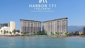 Ett modernt hotell med en framträdande skylt som visar texten ”Harbor 171 – Vallarta”. Hotellet består av en tvåvåningsbyggnad med balkonger och ligger vid en strand med palmer och en klar, blå himmel.