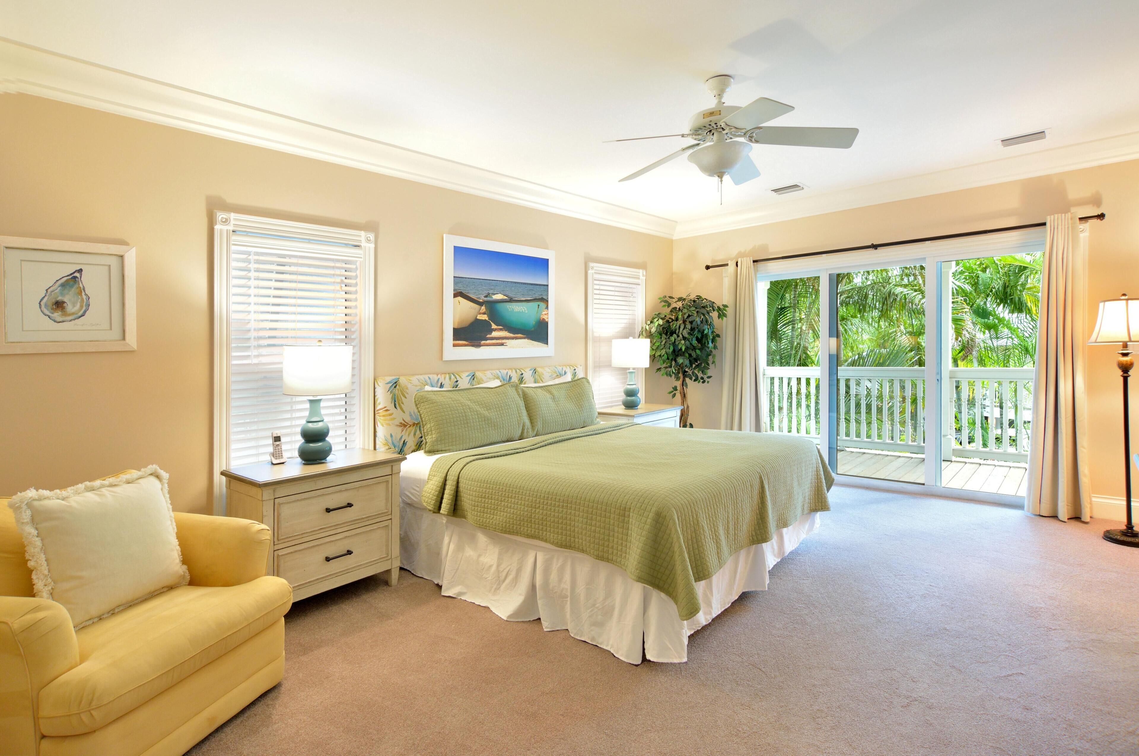 A bedroom with a bed, a nightstand, a lamp, a window with blinds, a ceiling fan, and a view of the outdoors.