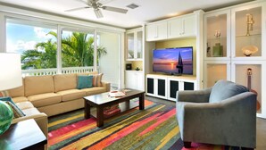 A living room with a sofa, armchair, coffee table, and TV. There is a ceiling fan, windows with a view of palm trees, and a colorful rug. - Island Chi c (Key West)
