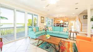 A spacious living room with a large teal sofa, a wooden coffee table, and a colorful rug. A balcony with a wooden railing overlooks a body of water. - Banana Breez e (Key West)