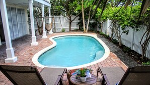 A small, curved swimming pool with a white edge, surrounded by a brick patio. There are two wooden chairs and a small round table with a plant and a magazine. - Banana Breez e (Key West)
