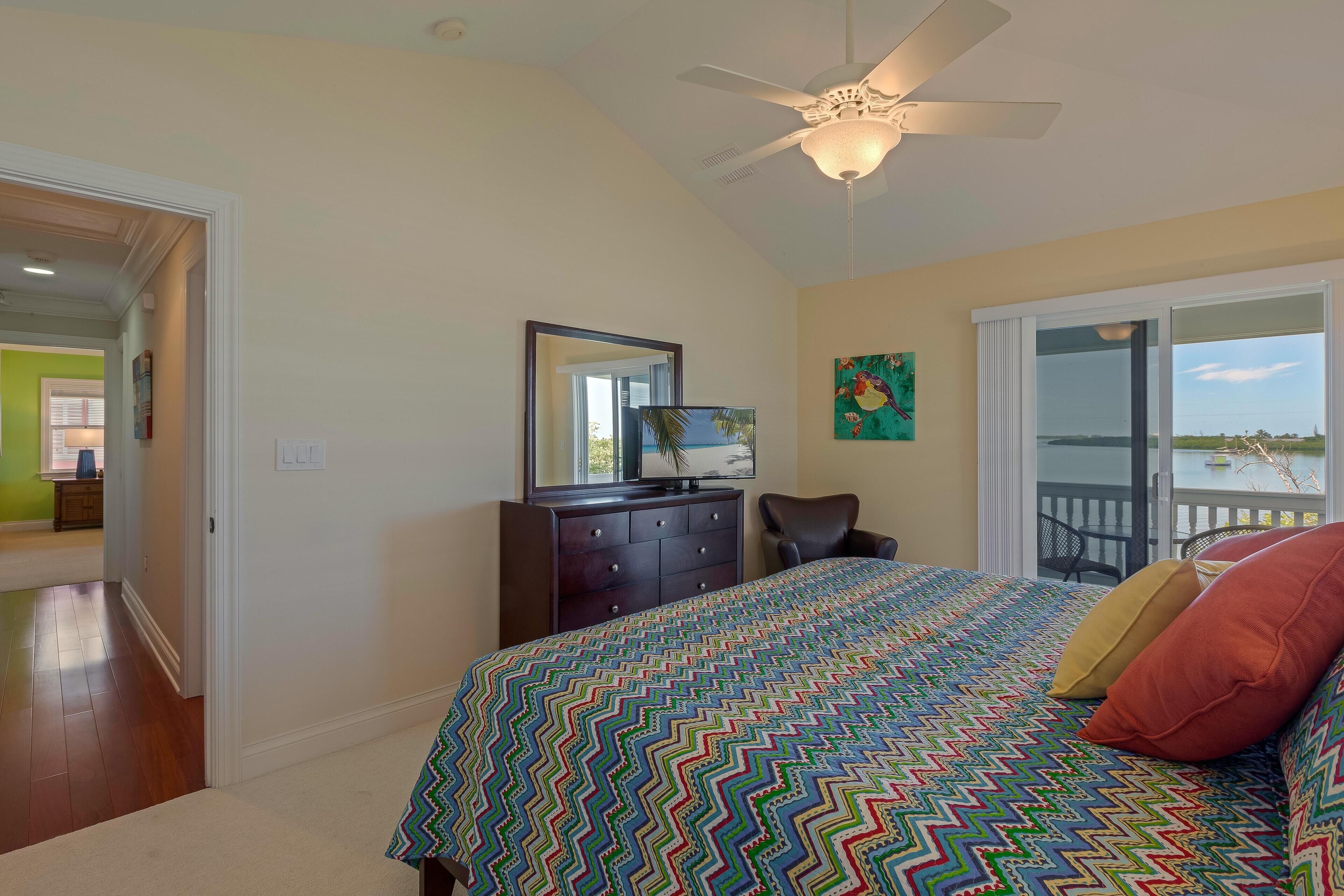 A bedroom with a bed, a dresser, a chair, a ceiling fan, and a view of the outdoors through a sliding door.