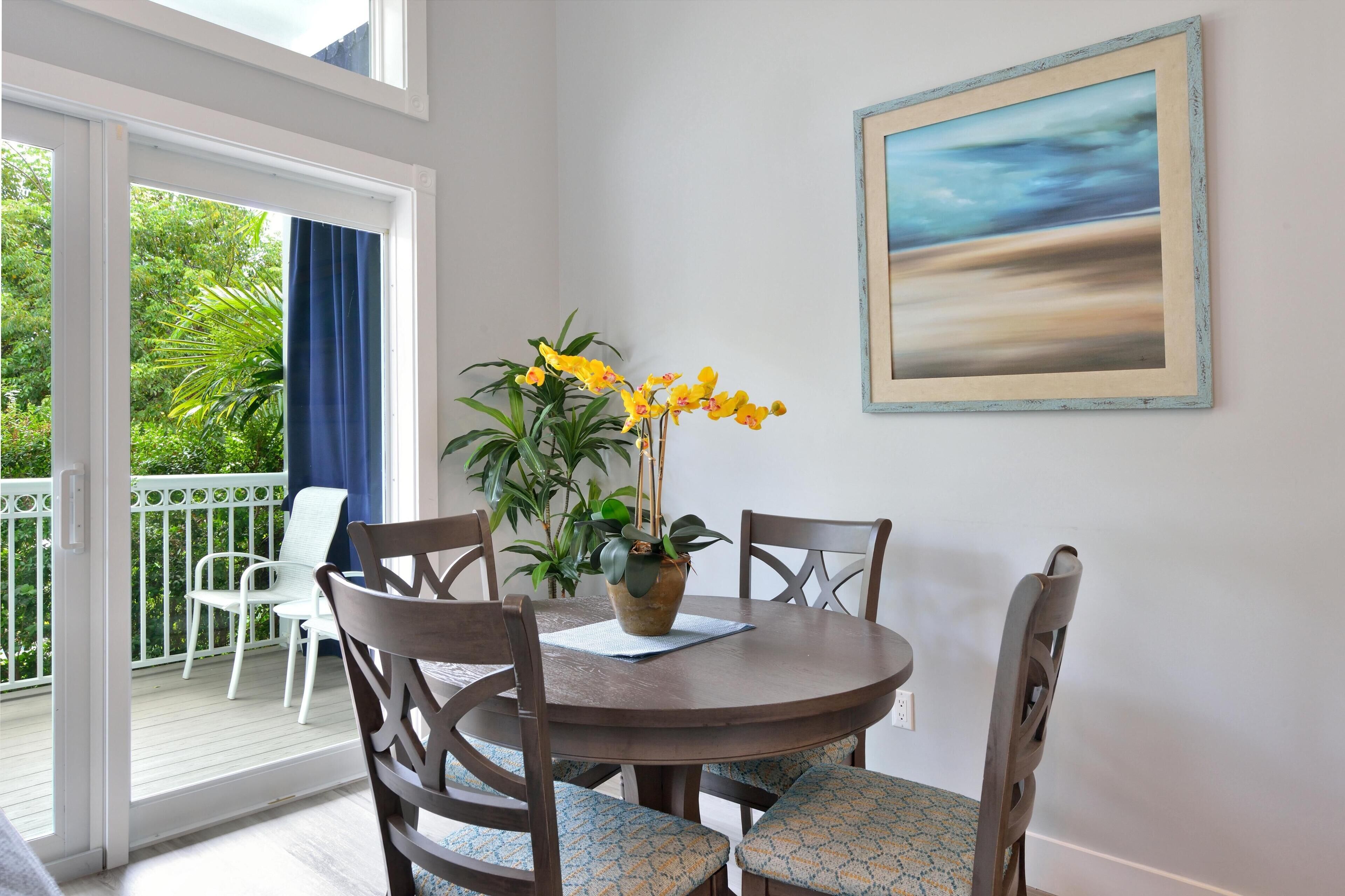 Un coin repas avec une table ronde, quatre chaises, une plante en pot aux fleurs jaunes et un tableau encadré accroché au mur.