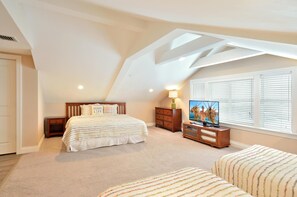 Quarto com teto inclinado, cama com roupa de cama listrada, televisão sobre um suporte de madeira e janela com persianas.