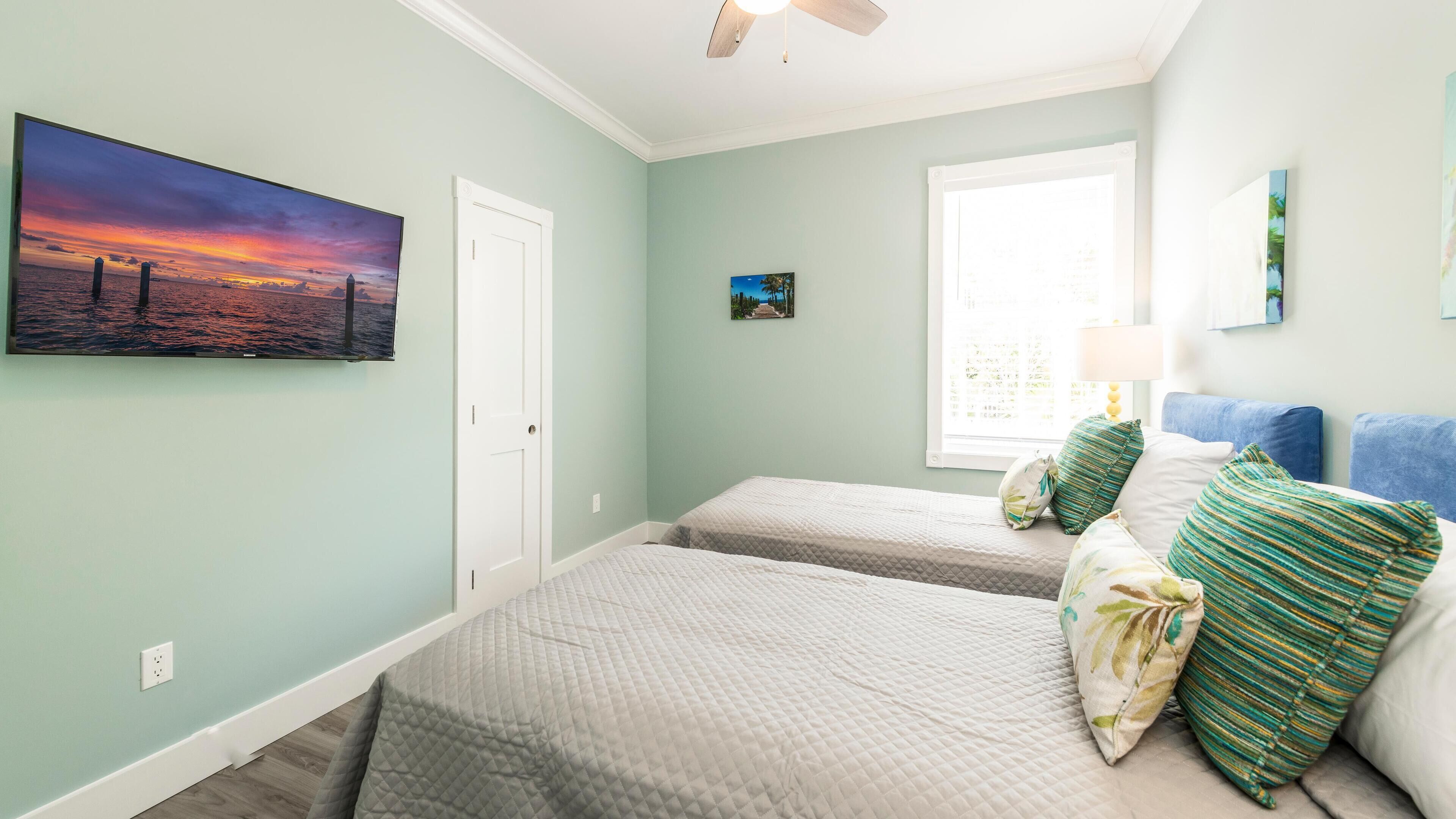 A bedroom with two beds, a window with a view, a ceiling fan, and artwork on the walls.